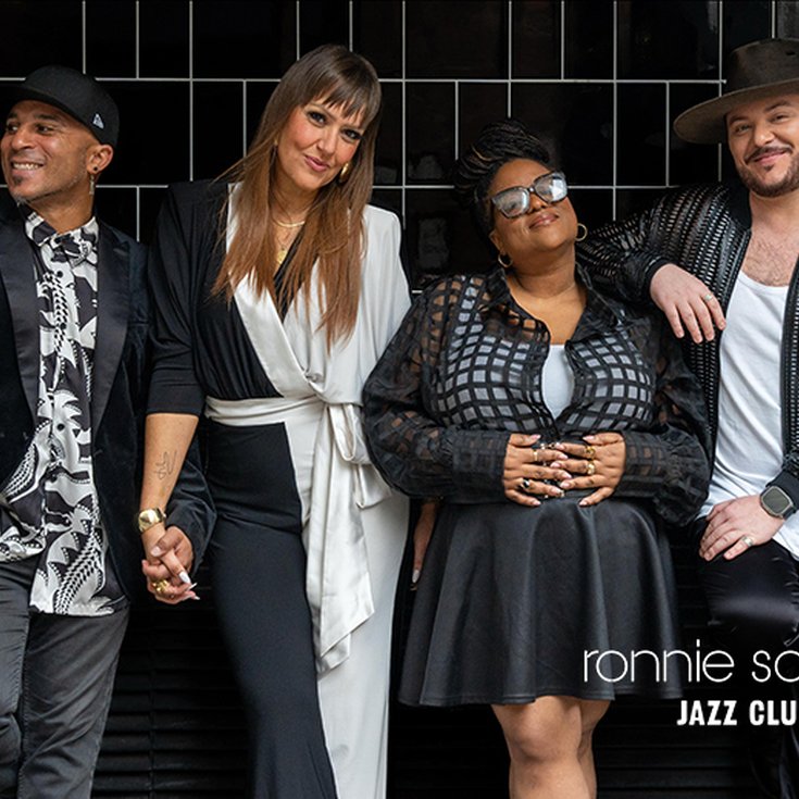 two females and two male singers all wearing black and white clothes standing against a black tiled wall