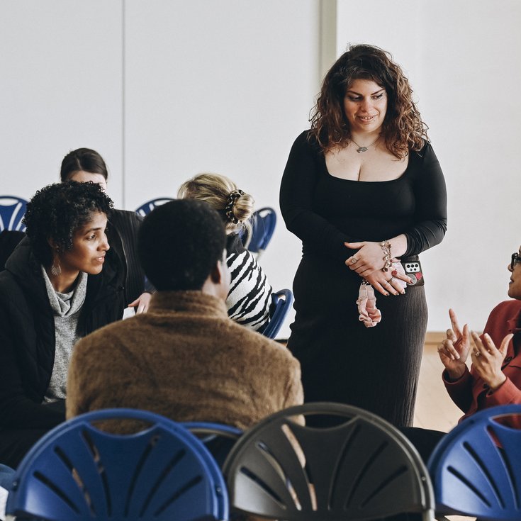 Small group discussion during a workshop or community event, with participants seated in a circle and engaged in conversation. One woman stands listening while another gestures as she speaks.