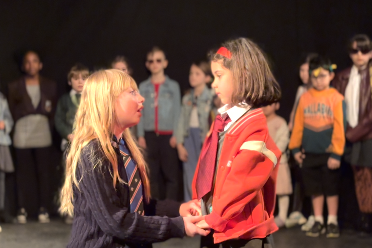 Two students sing while holding hands on stage as other students look on.