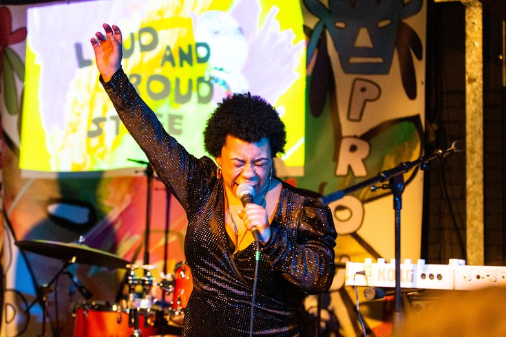 Black woman with short black hair wearing a black sparkly dress. She is singing into a mic with her right hand in the air and eyes closed.