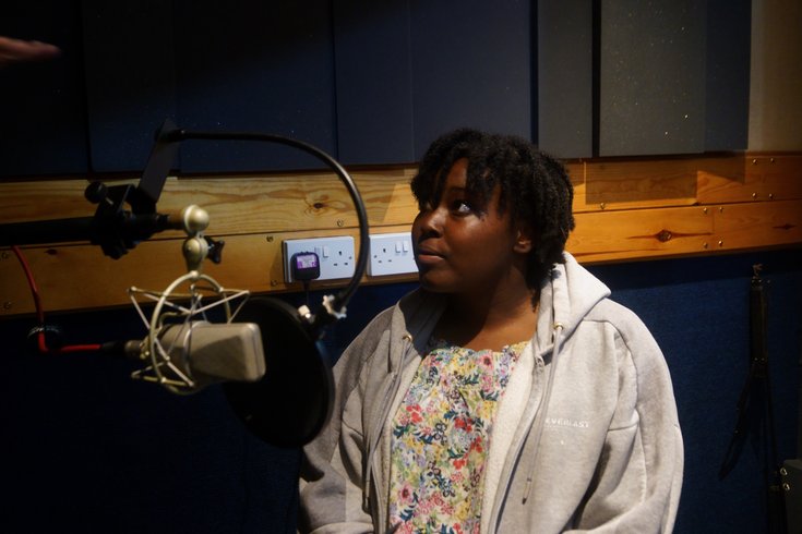 A young lady wearing a grey jumper over a floral top stands in a recording studio with a microphone in front of her.