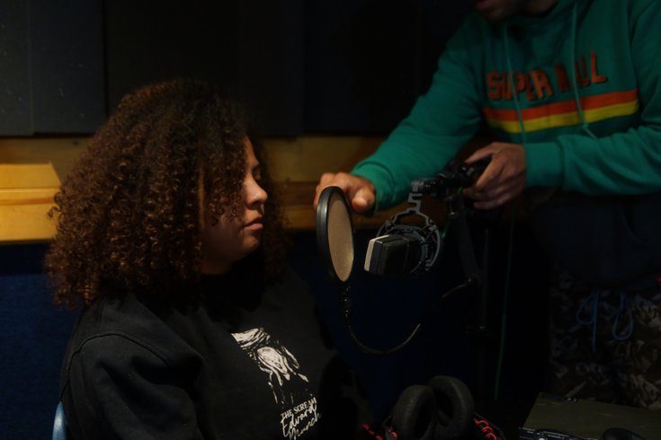 Harriet, with curly dark brown hair, prepares to record in The Midi Music Company studio for the 77 Watts podcast, as hands adjust the mic in front of her.