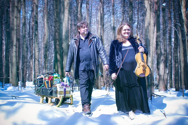 A scene of a forest with snow on the ground. A man pulls a sleigh and a woman holds a fiddle