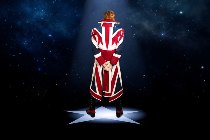 Person in a Union Jack coat stands on a star-shaped spotlight, against a starry night sky, hands clasped