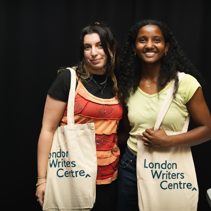 Two young women smile to camera carrying beige tote bags with the words London Writers Centre engraved on them.