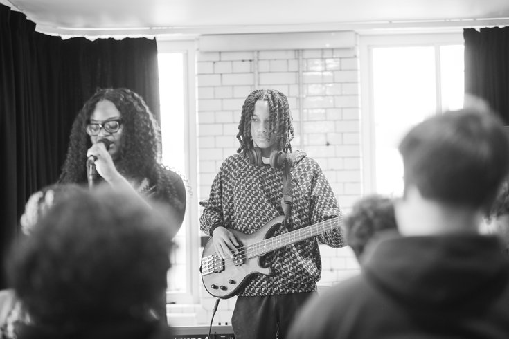 A young person sings into a mic in the foreground and another plays the bass guitar in the background.
